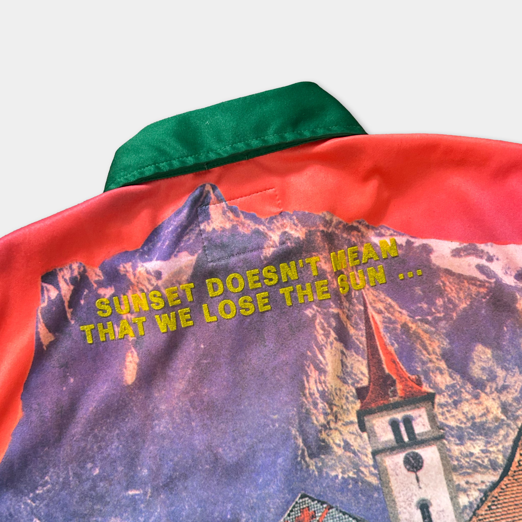 a colorful jacket with a printed design featuring a mountain landscape and a church steeple. The text on the jacket reads "SUNSET DOESN'T MEAN THAT WE LOSE THE SUN..."
