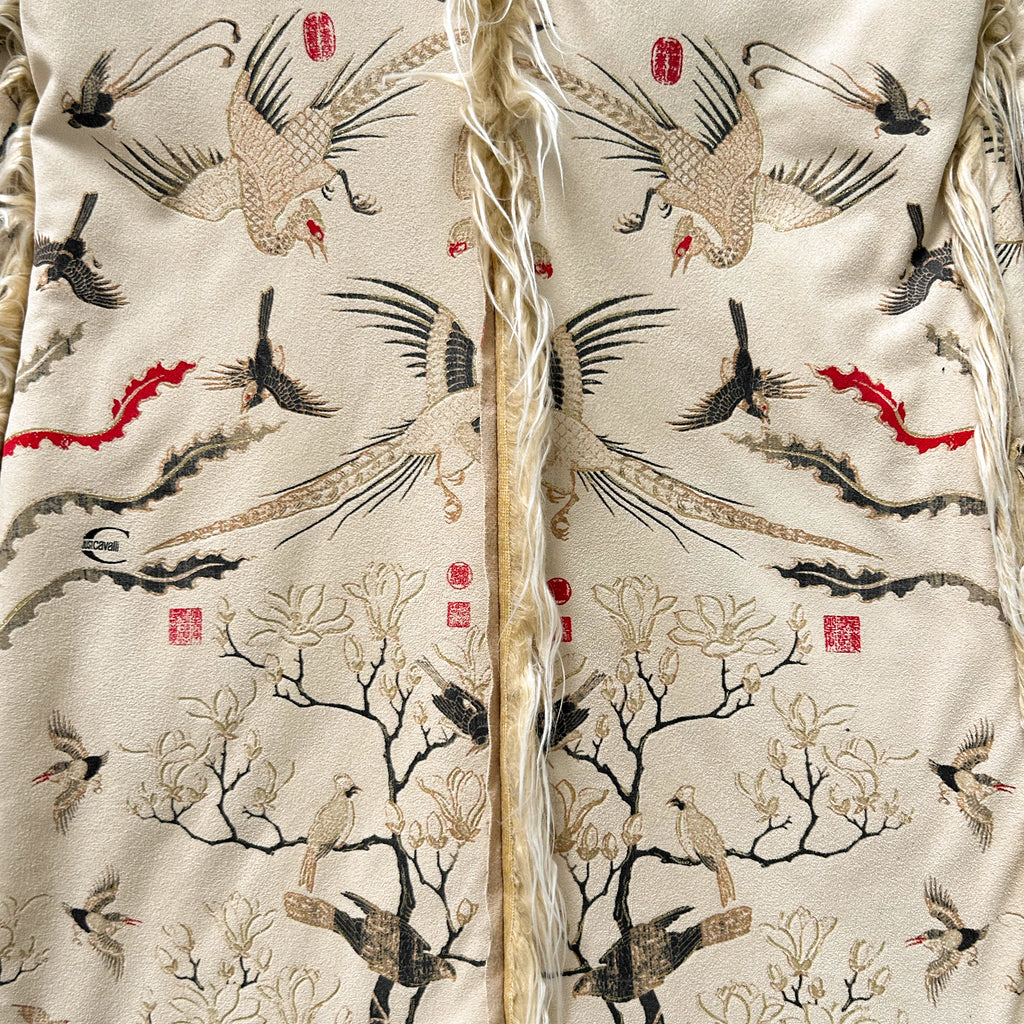 a close-up view of a fabric with a repeating pattern of birds and trees, including a large bird with a long neck and a smaller bird with a shorter neck. The fabric appears to be a beige material with a fringe at the bottom.