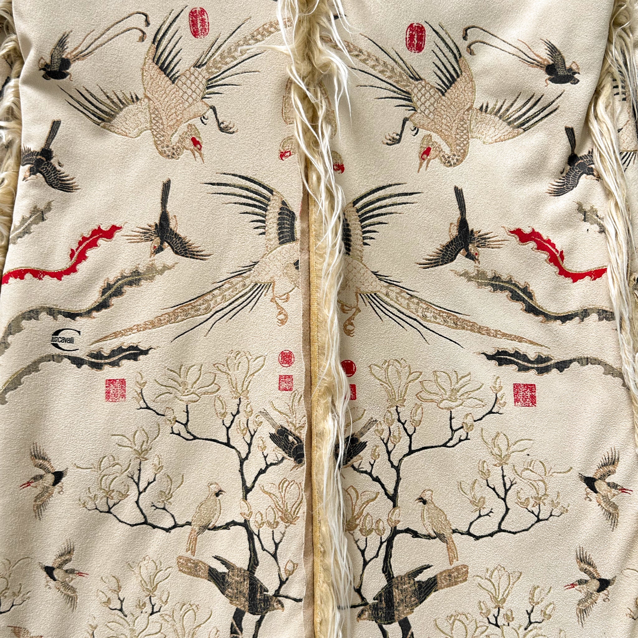 a close-up view of a fabric with a repeating pattern of birds and trees, including a large bird with a long neck and a smaller bird with a shorter neck. The fabric appears to be a beige material with a fringe at the bottom.
