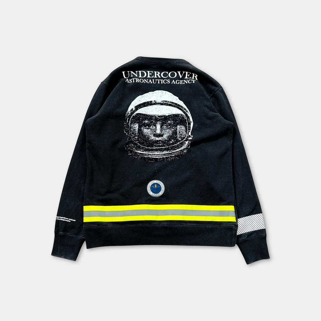 a black sweatshirt with a graphic of a person wearing a spacesuit and the text "Undercover Astronautics Agency" printed on the back.