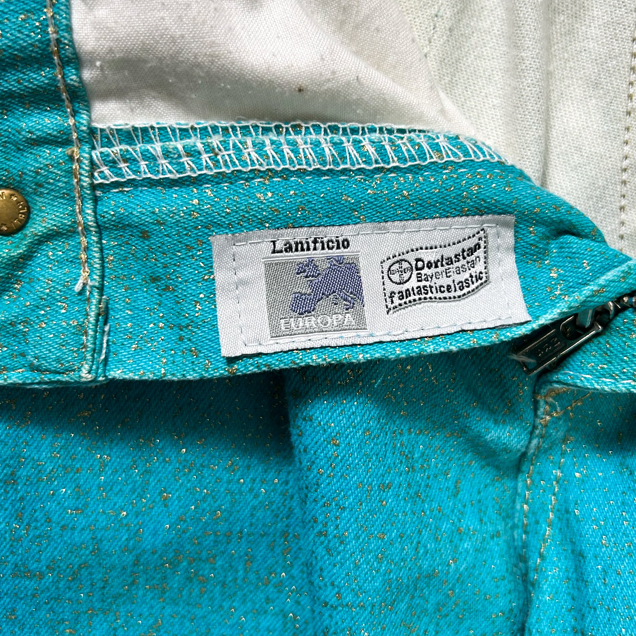 A close-up view of a teal-colored denim fabric, with a label attached to it that reads "Lanzifico" and "Dorostan Fabrics fantastic gesticulate".