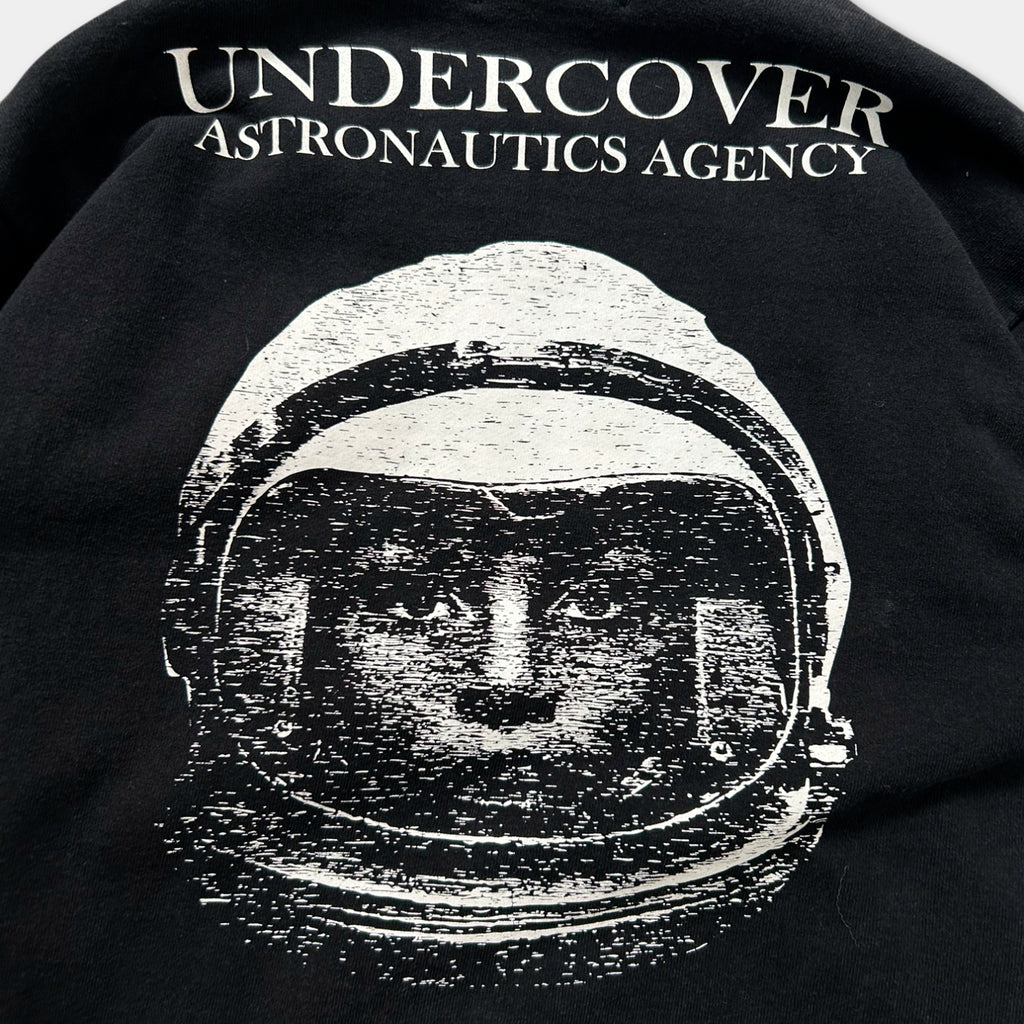 a black and white graphic of a space helmet with the text "Undercover Astronautics Agency" printed on the back of a black shirt.