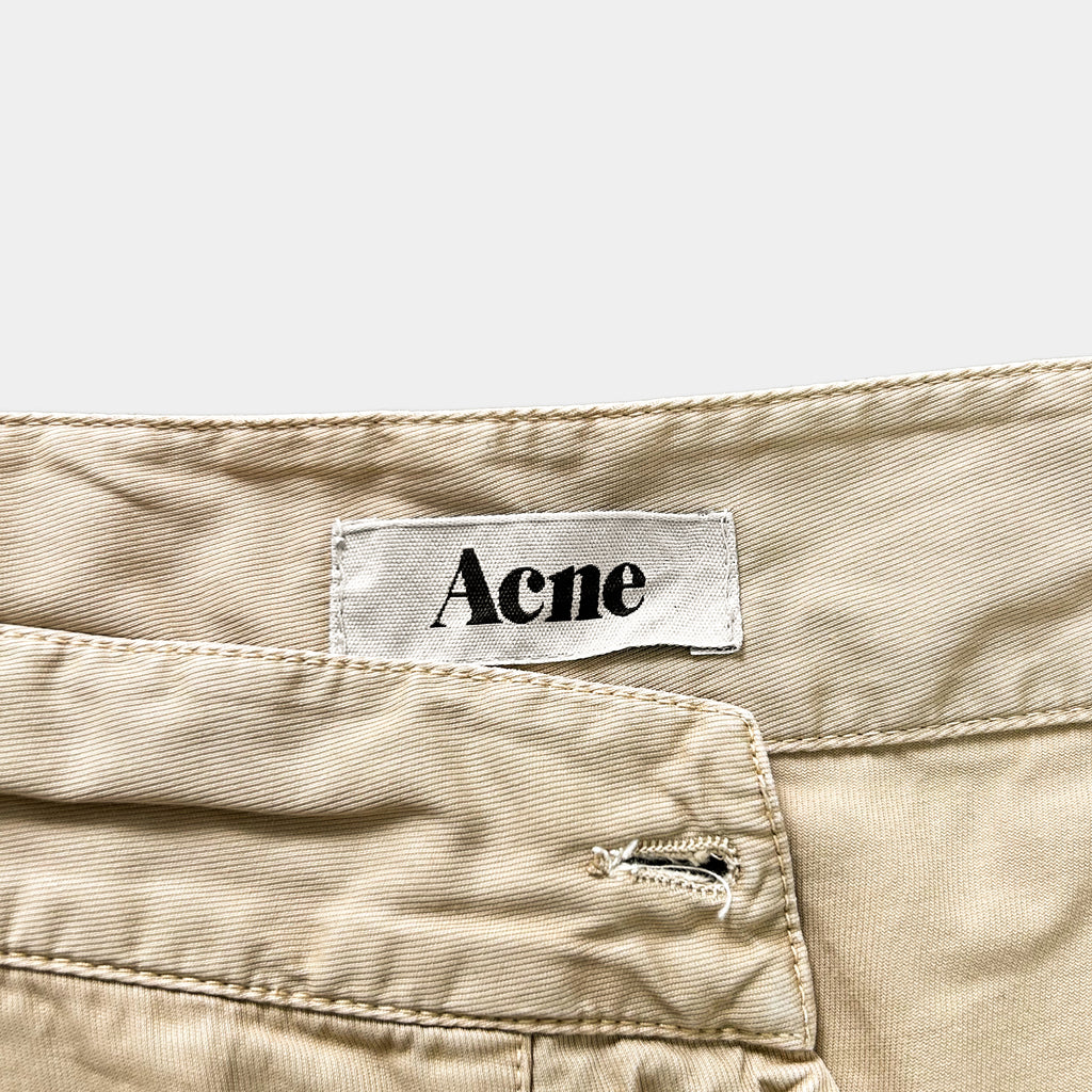 A label with the word "Acne" printed on it is attached to a beige fabric, which appears to be a pair of pants or shorts.