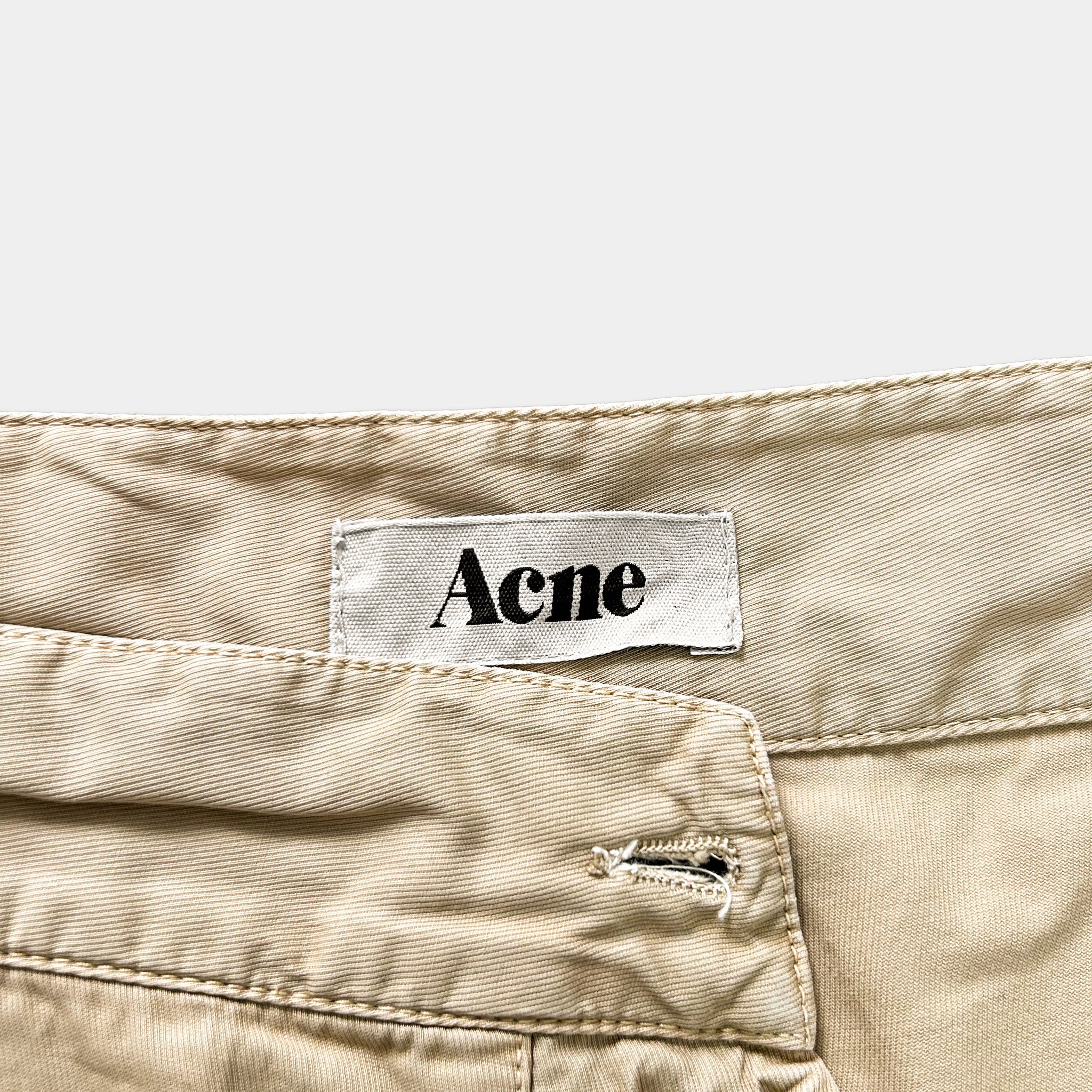 A label with the word "Acne" printed on it is attached to a beige fabric, which appears to be a pair of pants or shorts.