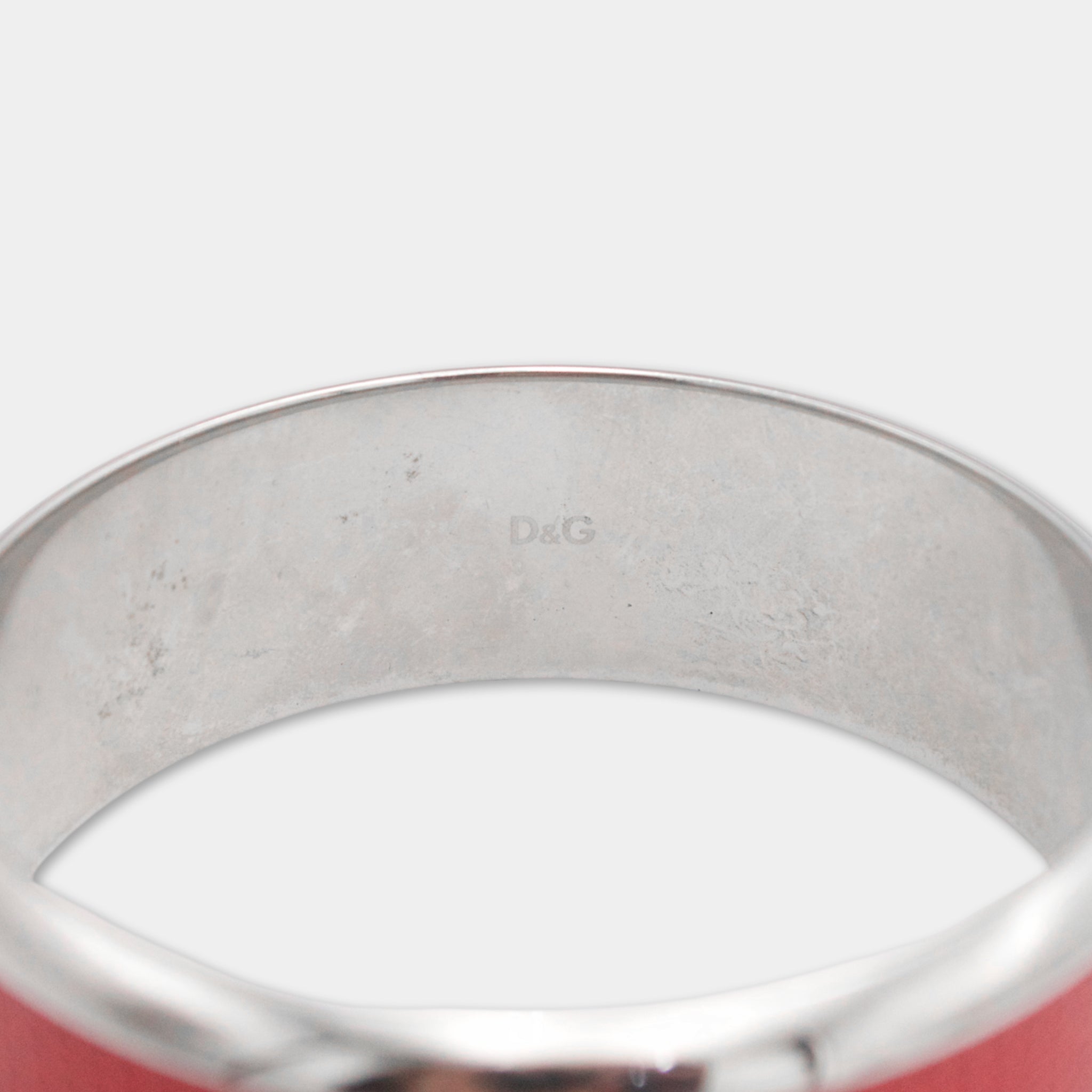 A close-up view of a silver bracelet with a red band, showing the engraved logo of the luxury brand D'G.