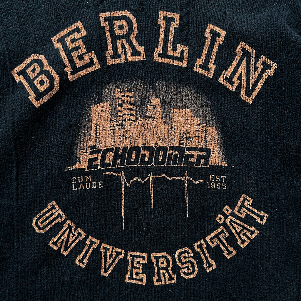 a close-up of a dark-colored fabric with text and graphics printed on it. The text includes the words "BERLIN", "ECHODOMER", "SUM LAUDÉ", "UNIVERSITÄT", and "EST 1995". The graphics depict a cityscape with buildings and a heart shape. The text and graphics are arranged in a circular pattern, creating a visually appealing design.