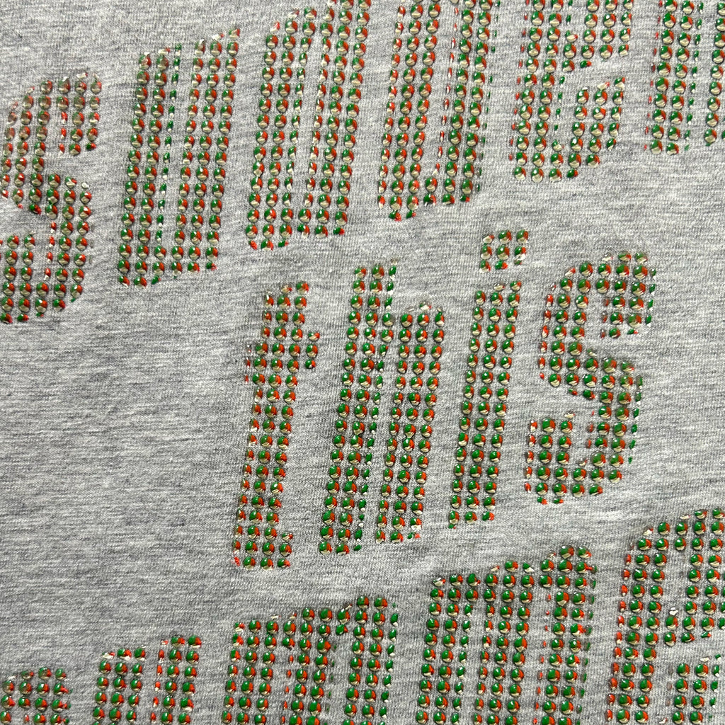 a close-up view of a grey fabric with a pattern of red and green dots or beads arranged in a repeating design. The pattern is not uniform, with some areas having more red dots than green ones, and the overall design is not symmetrical. The image does not contain any text or other discernible objects.