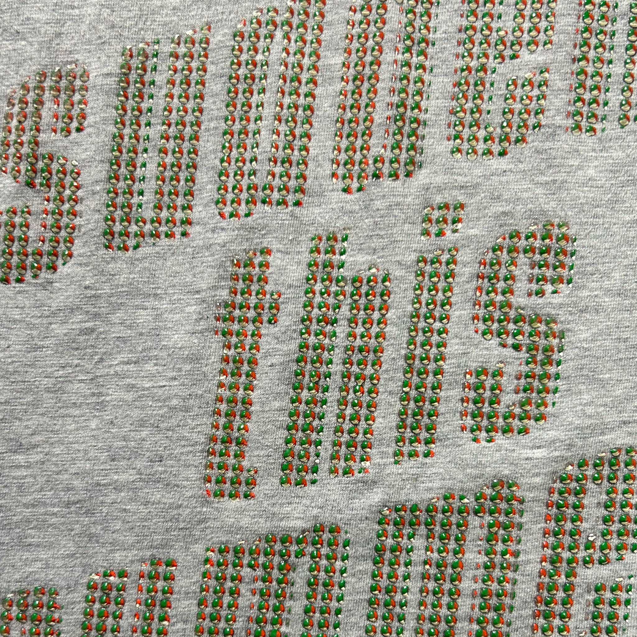 a close-up view of a grey fabric with a pattern of red and green dots or beads arranged in a repeating design. The pattern is not uniform, with some areas having more red dots than green ones, and the overall design is not symmetrical. The image does not contain any text or other discernible objects.