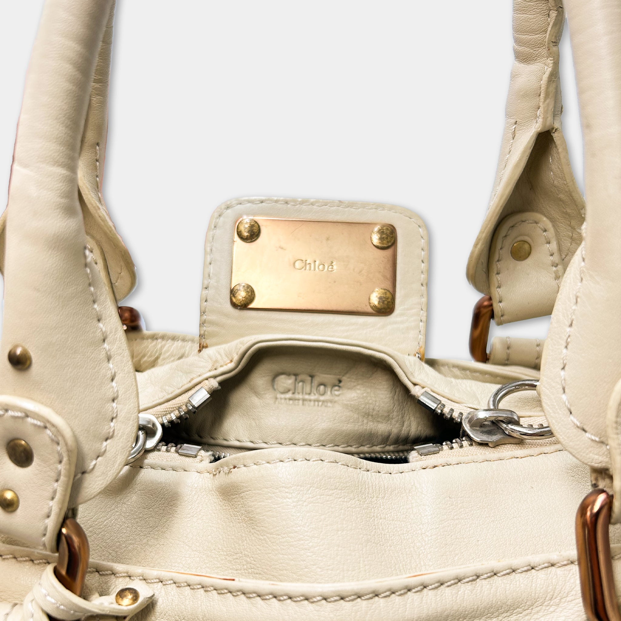 A close-up view of a beige leather handbag with a gold-colored metal plate attached to the front.