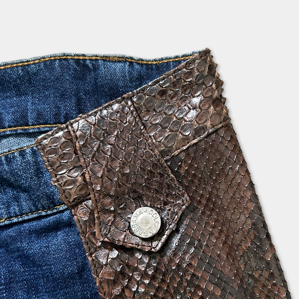 A close-up view of a brown leather wallet or card case with a snake skin texture, placed on top of a pair of blue denim jeans.
