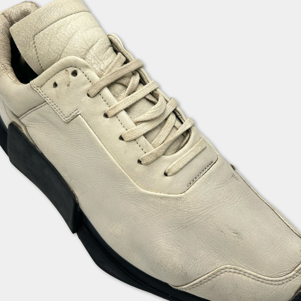 A close-up view of a white leather sneaker with a black sole, showing the upper part of the shoe and the laces.