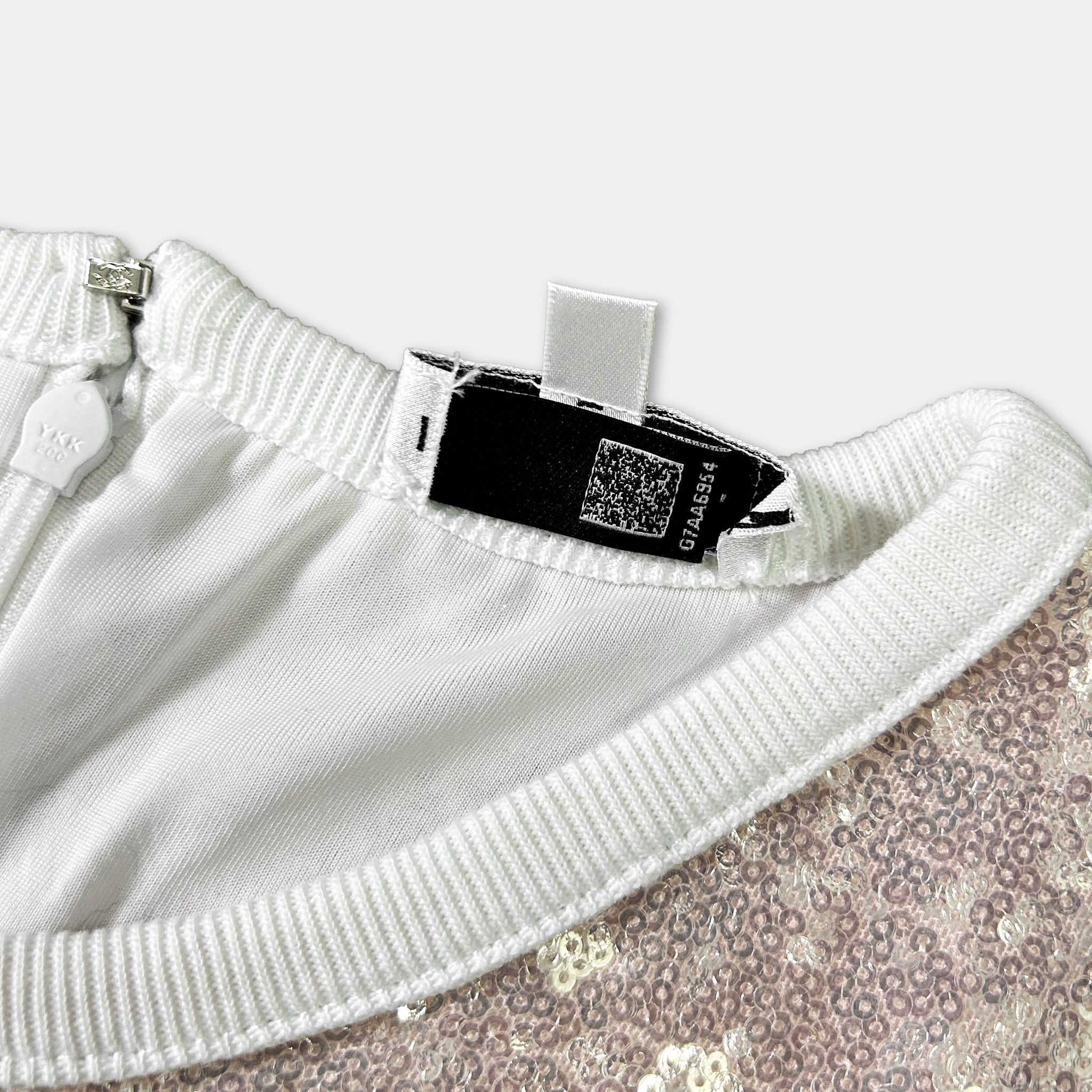 a close-up view of a white garment with a sequin pattern, featuring a tag attached to the collar.