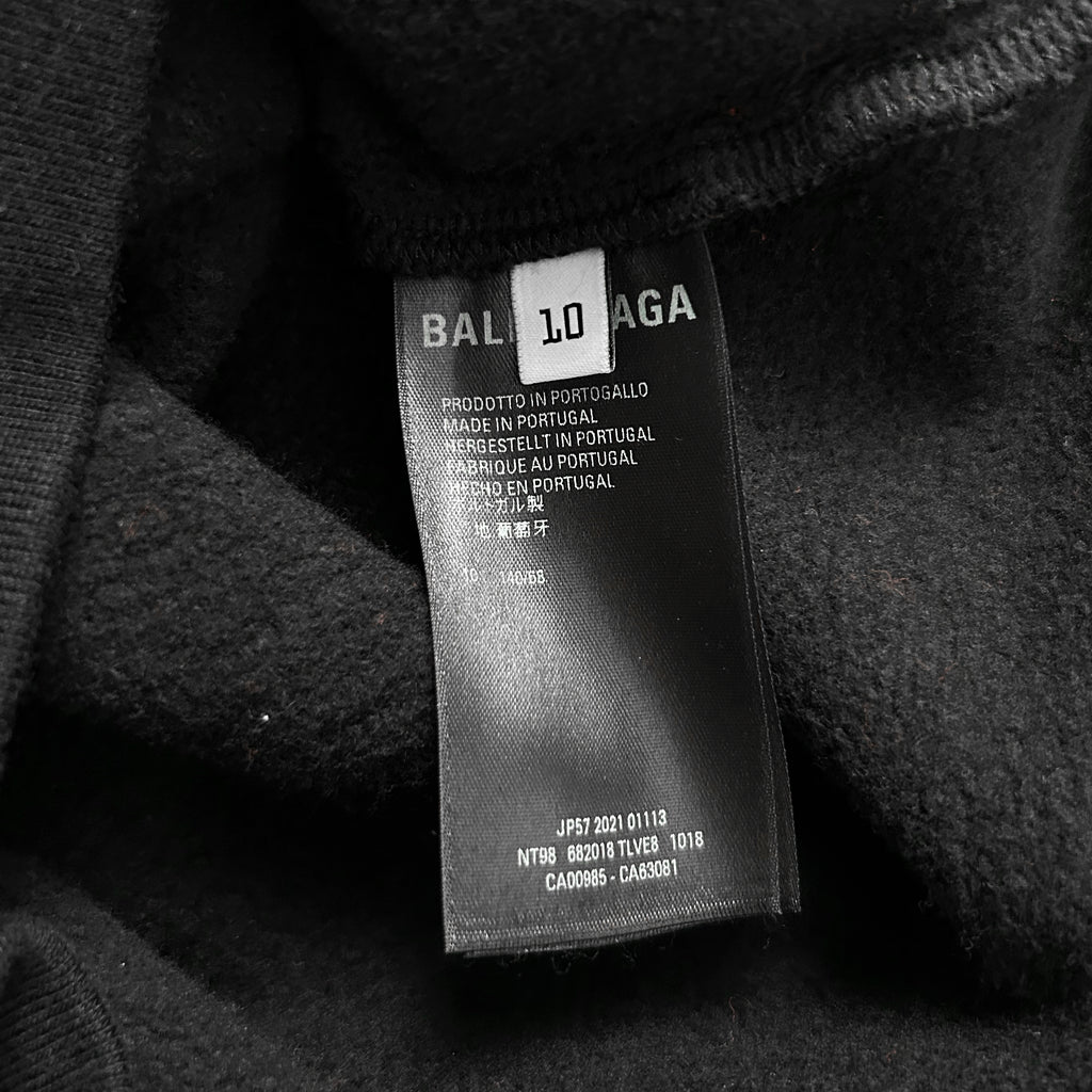 a close-up view of a black fabric, possibly a sweater or garment, with a label attached to it. The label appears to be a product label from Balenciaga, indicating that it is made in Portugal and is a T-shirt. The label also includes some text in a foreign language, which may be the country of origin or the manufacturer's name. The label is slightly wrinkled, suggesting that it has been worn or used before.