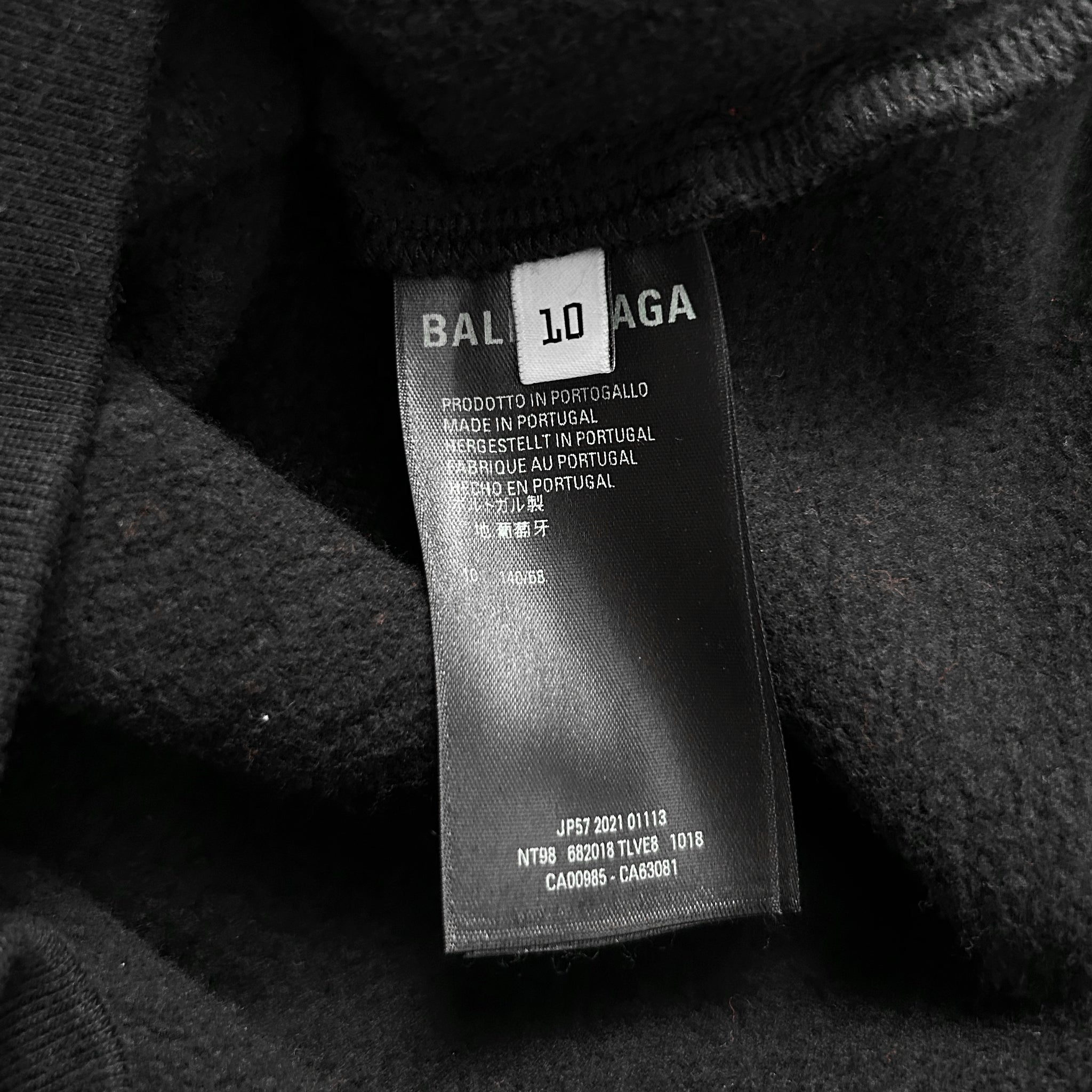 a close-up view of a black fabric, possibly a sweater or garment, with a label attached to it. The label appears to be a product label from Balenciaga, indicating that it is made in Portugal and is a T-shirt. The label also includes some text in a foreign language, which may be the country of origin or the manufacturer's name. The label is slightly wrinkled, suggesting that it has been worn or used before.