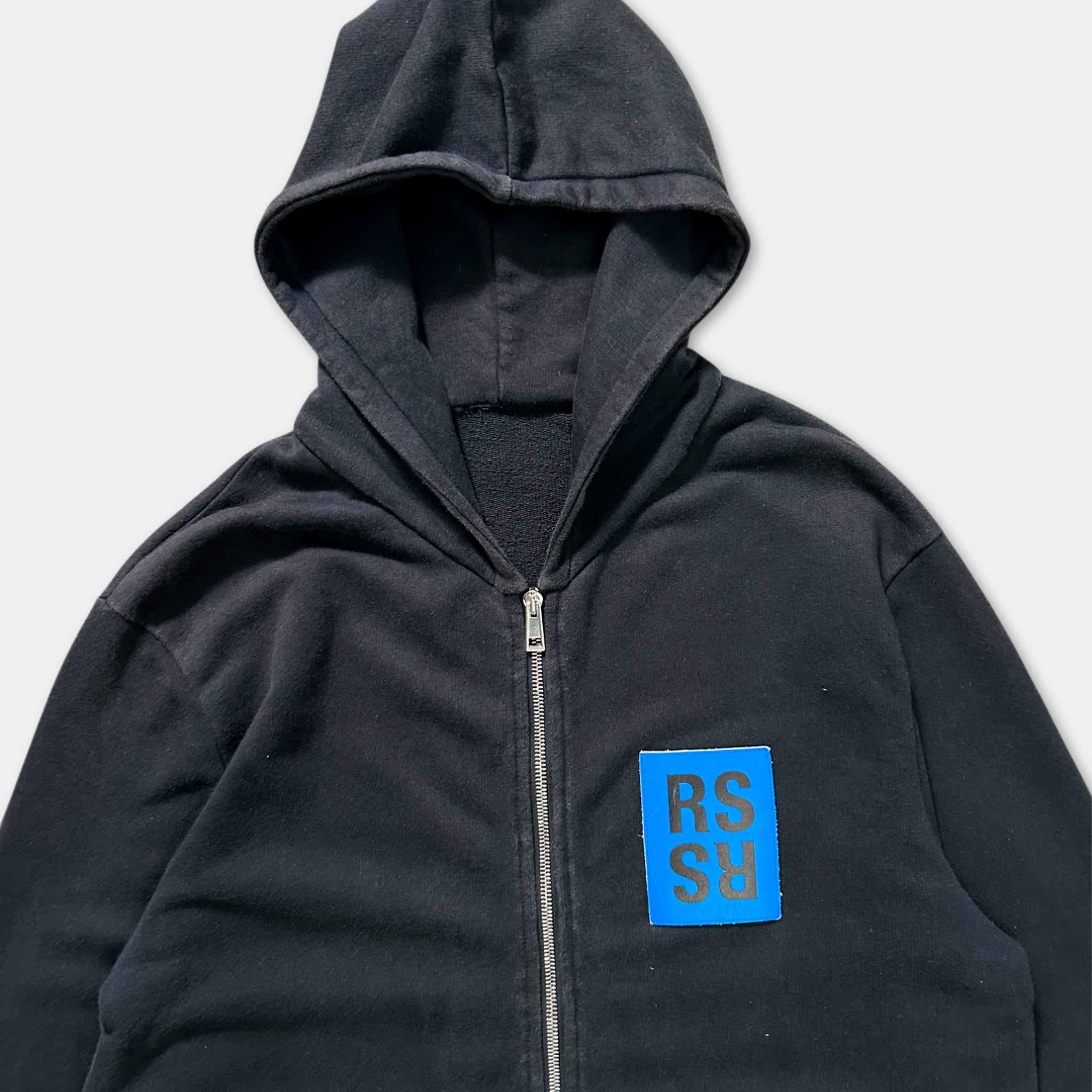 A black zip-up hoodie with a blue patch on the front displaying the letters 'RS' in blue.