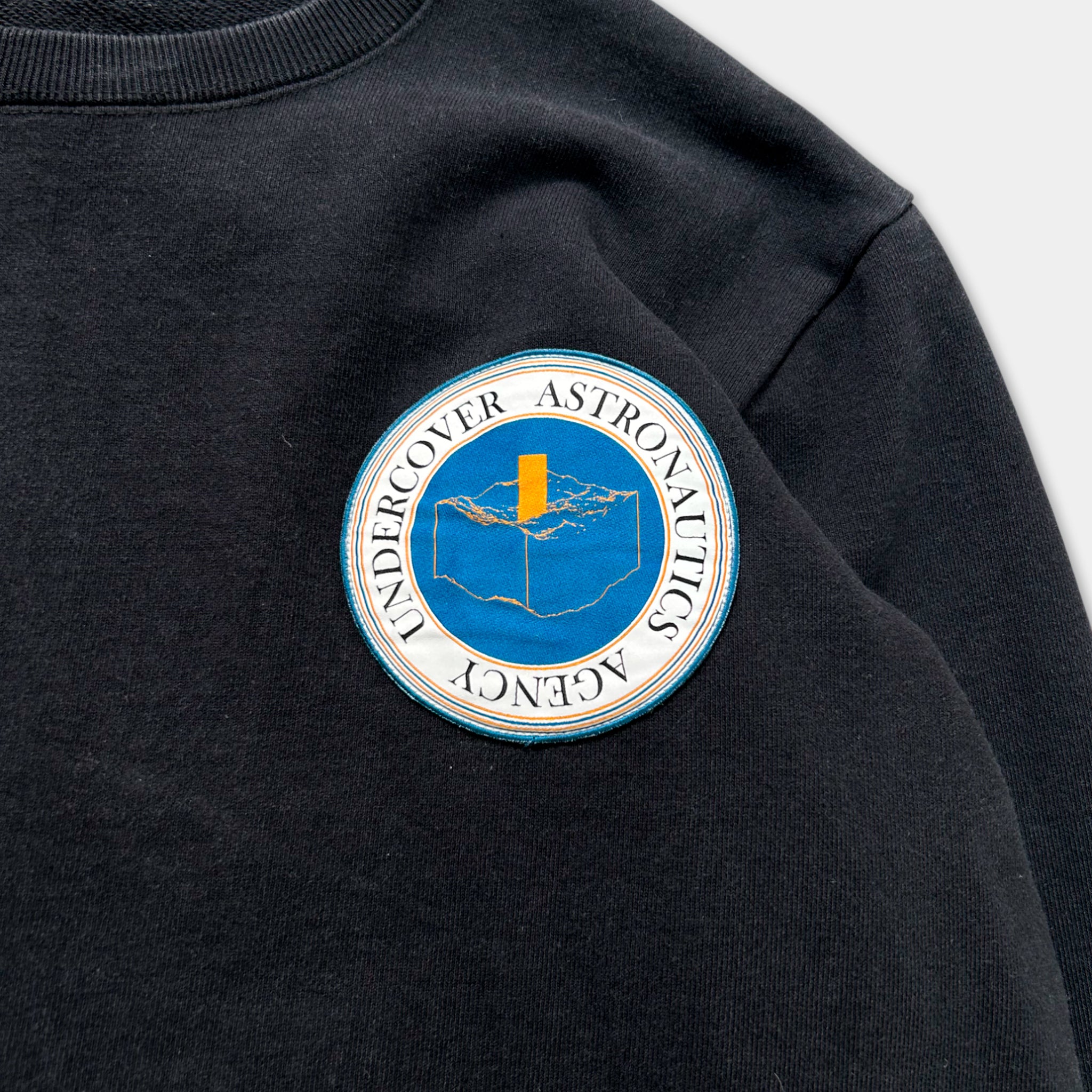 a black sweater with a circular patch on the front, featuring a blue and yellow design and the text "Undercover Astronautics".