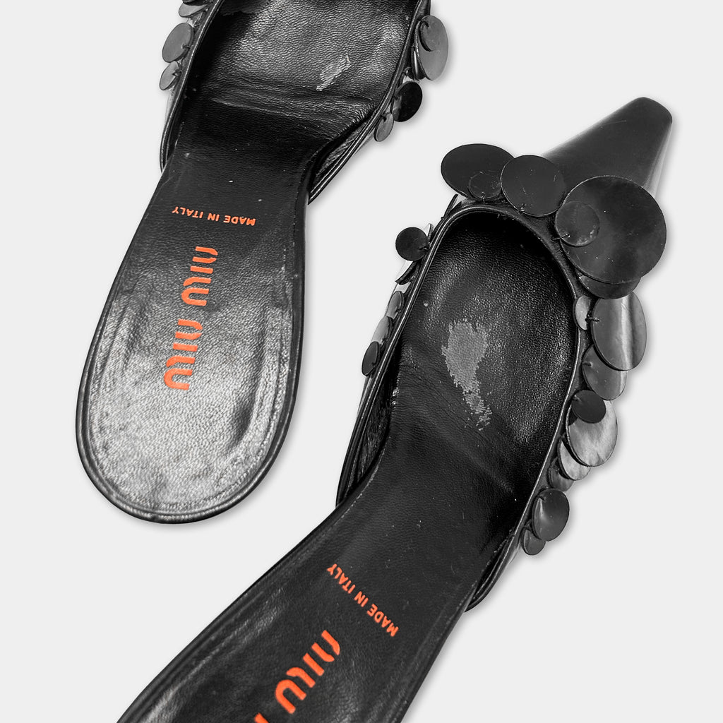 A pair of black high-heeled shoes with a distinctive design, featuring a pointed toe and circular embellishments.