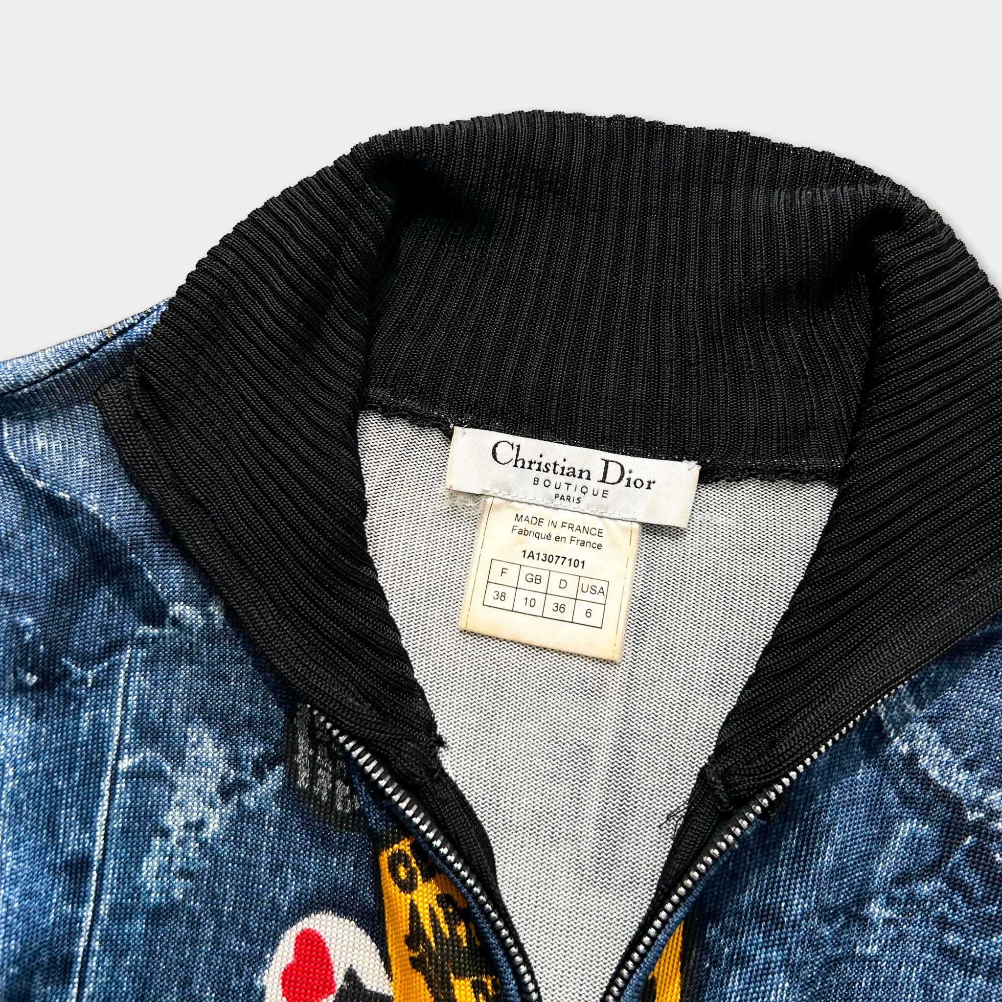 A close-up view of a blue denim jacket with a black collar and zipper, featuring a label that reads "Christian Dior" and "Made in France Fabrics France/USA".