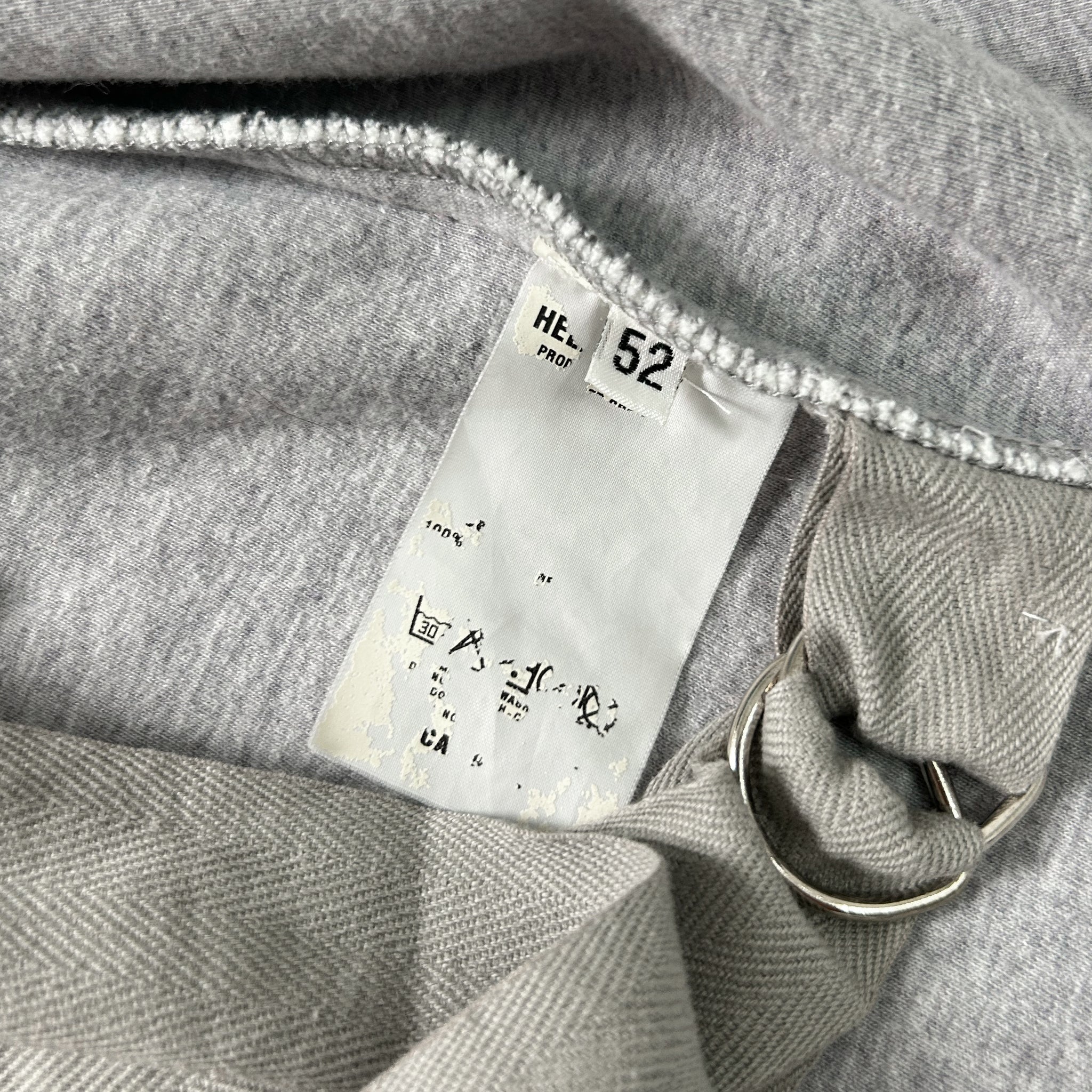 a close-up view of a grey fabric, possibly a sweater or garment, with a white label attached to it. The label appears to contain text and numbers, possibly indicating the product's details or measurements.