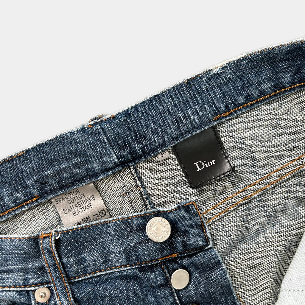 A close-up view of a pair of blue denim jeans, with a label attached to the waistband and a Dior logo visible on the waistband.