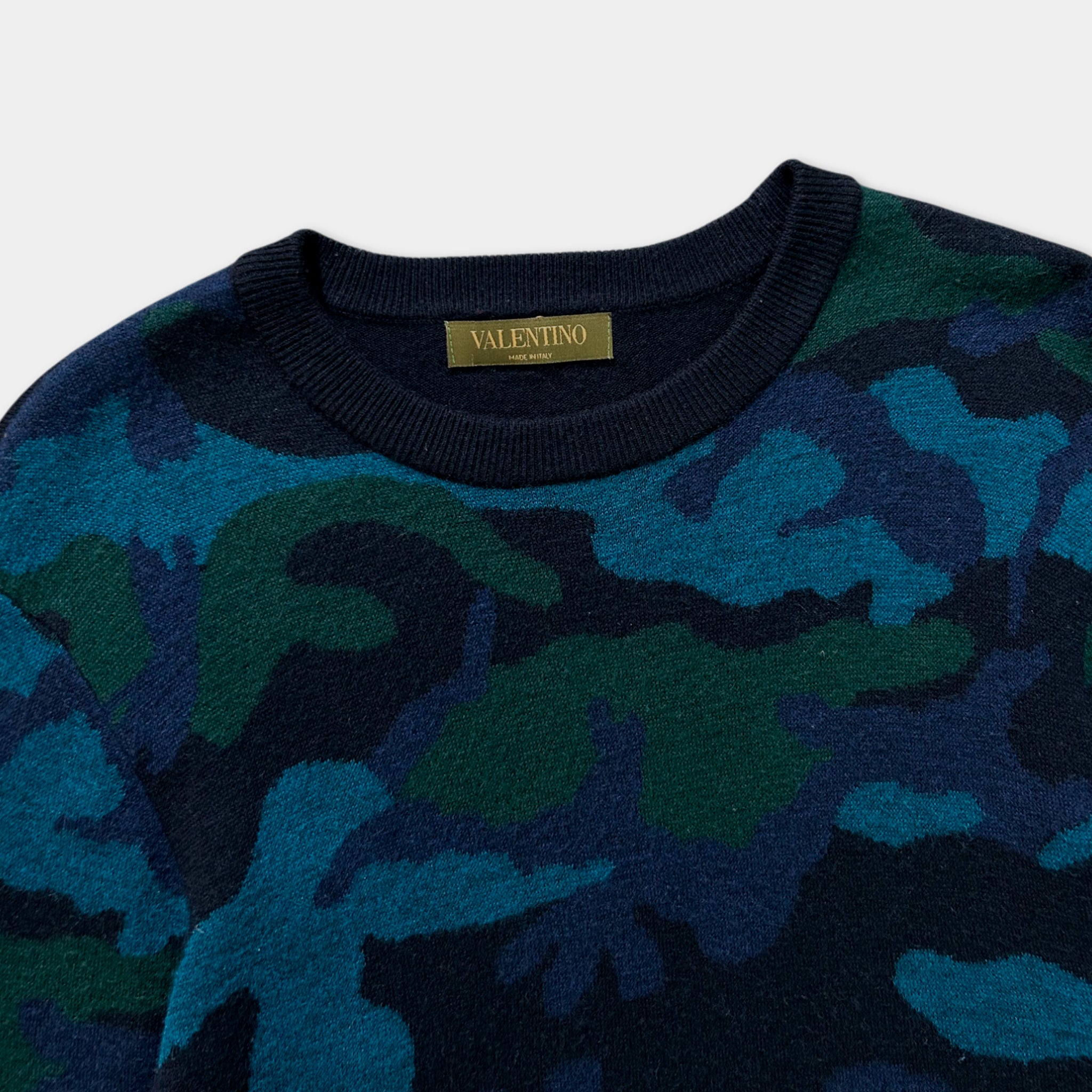 A close-up image of a blue and green camouflage-patterned sweater with a gold label on the collar.