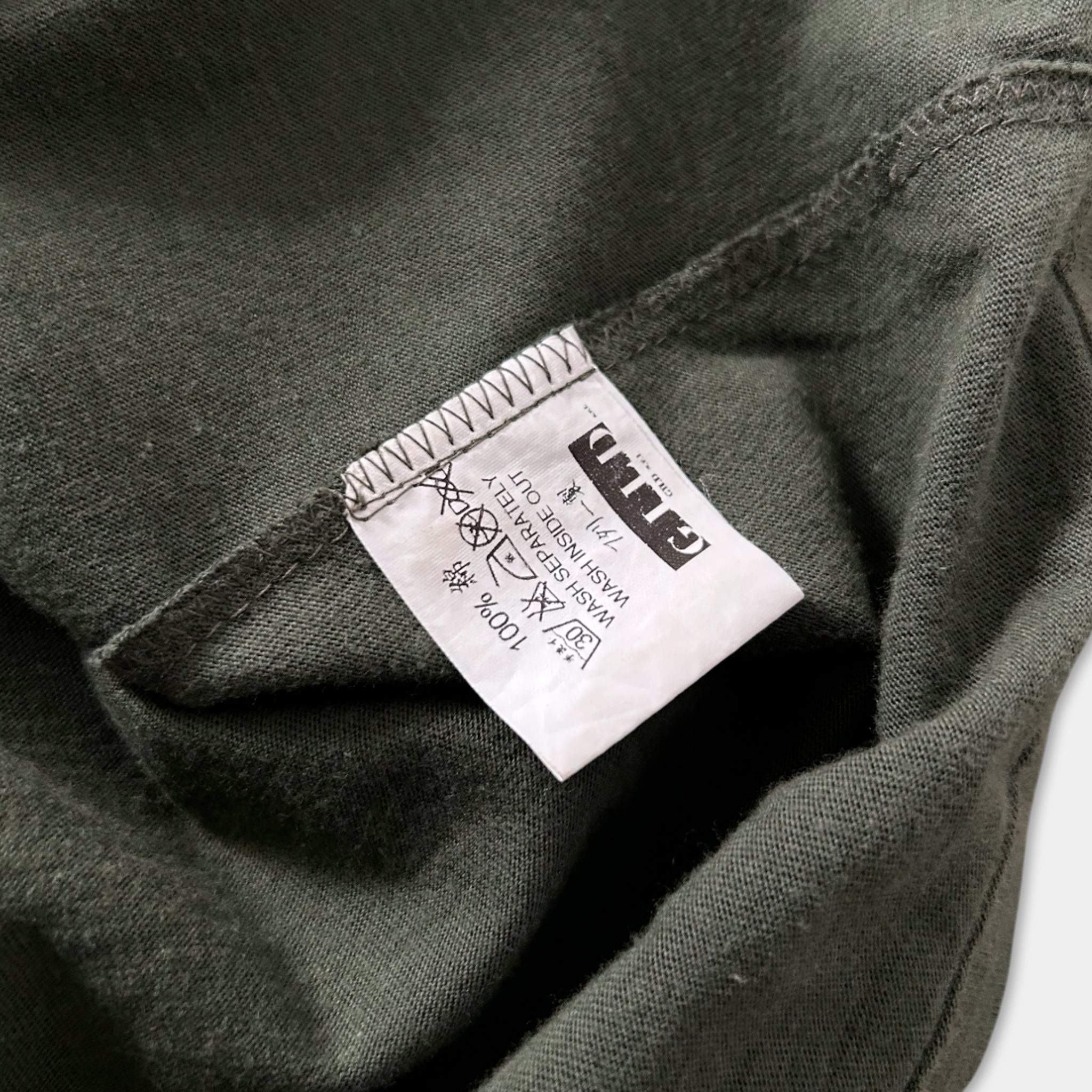 a close-up view of a dark green fabric, possibly a t-shirt or garment, with a white label attached to it. The label appears to contain text and symbols, but the exact wording is not clearly visible.