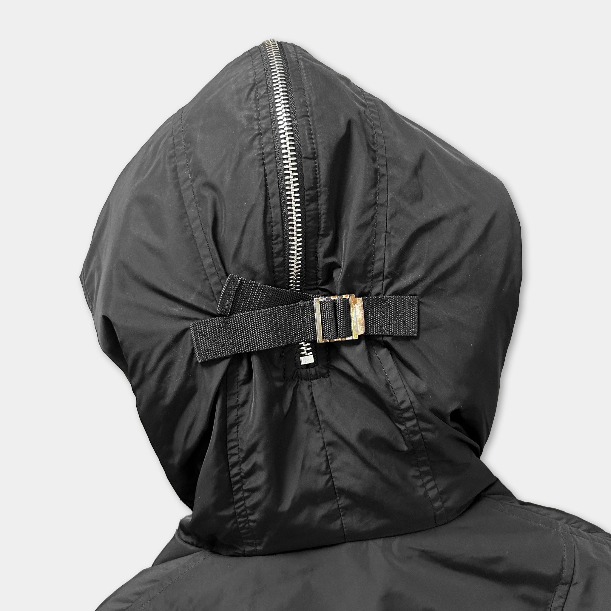 A close-up view of the back of a black hooded jacket with a zipper and a black strap.