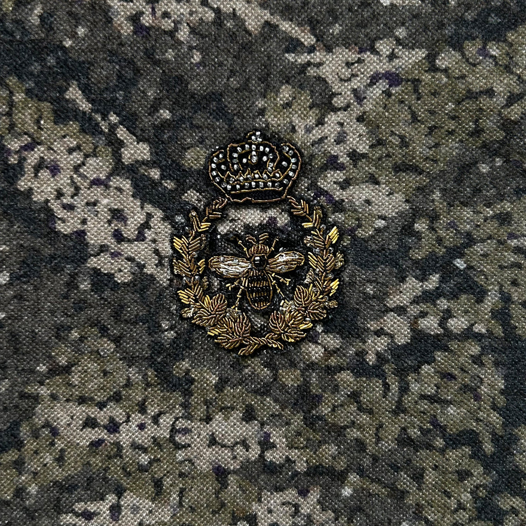 a decorative badge with a crown and a bee, set against a camouflage-like background.