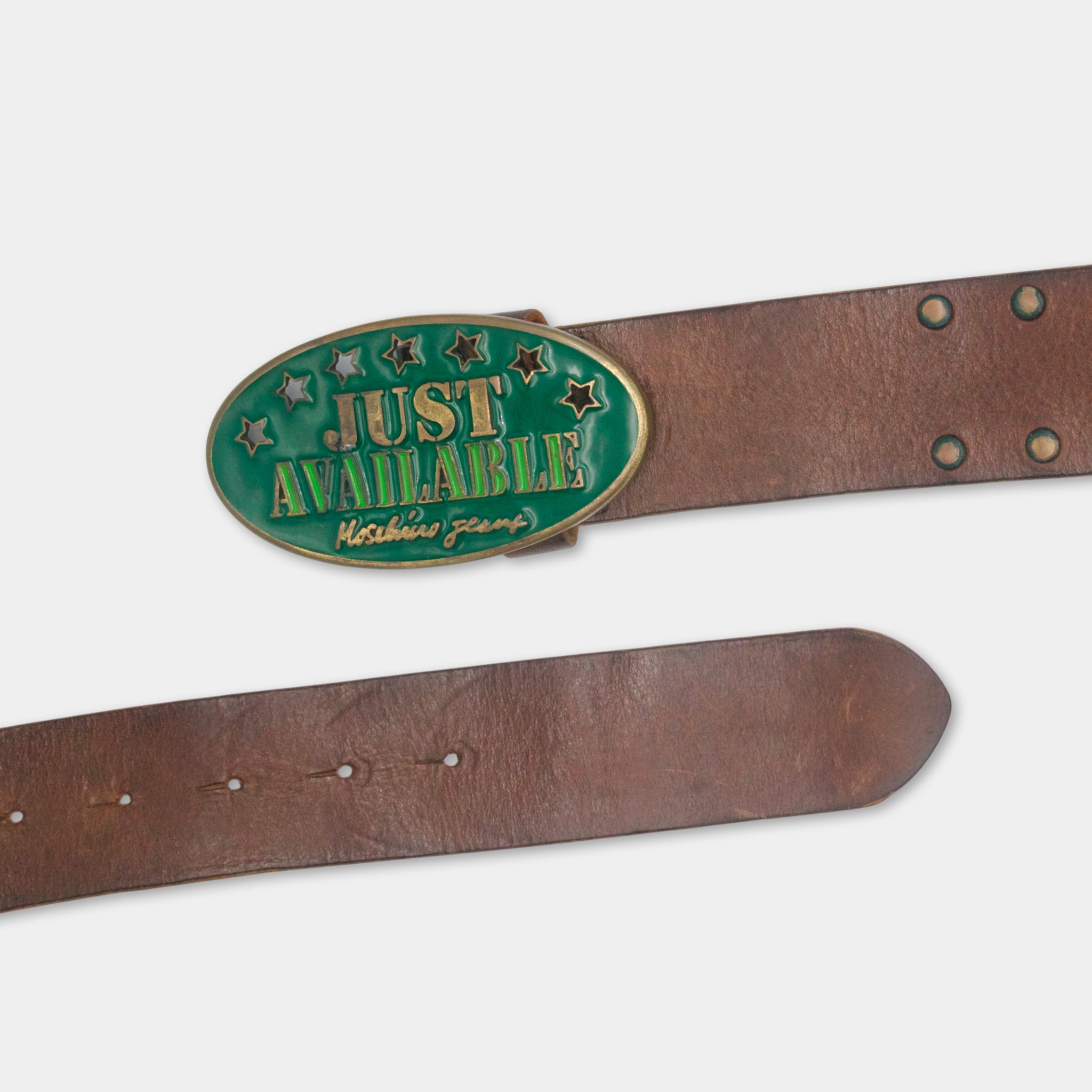 A brown leather belt with a green oval-shaped buckle that says "JUST AVAILABLE" and has stars on it.