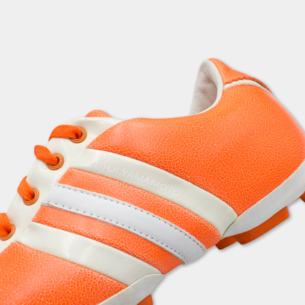 A close-up of an orange and white Adidas cleat with the brand name "Yamamoto" printed on it.
