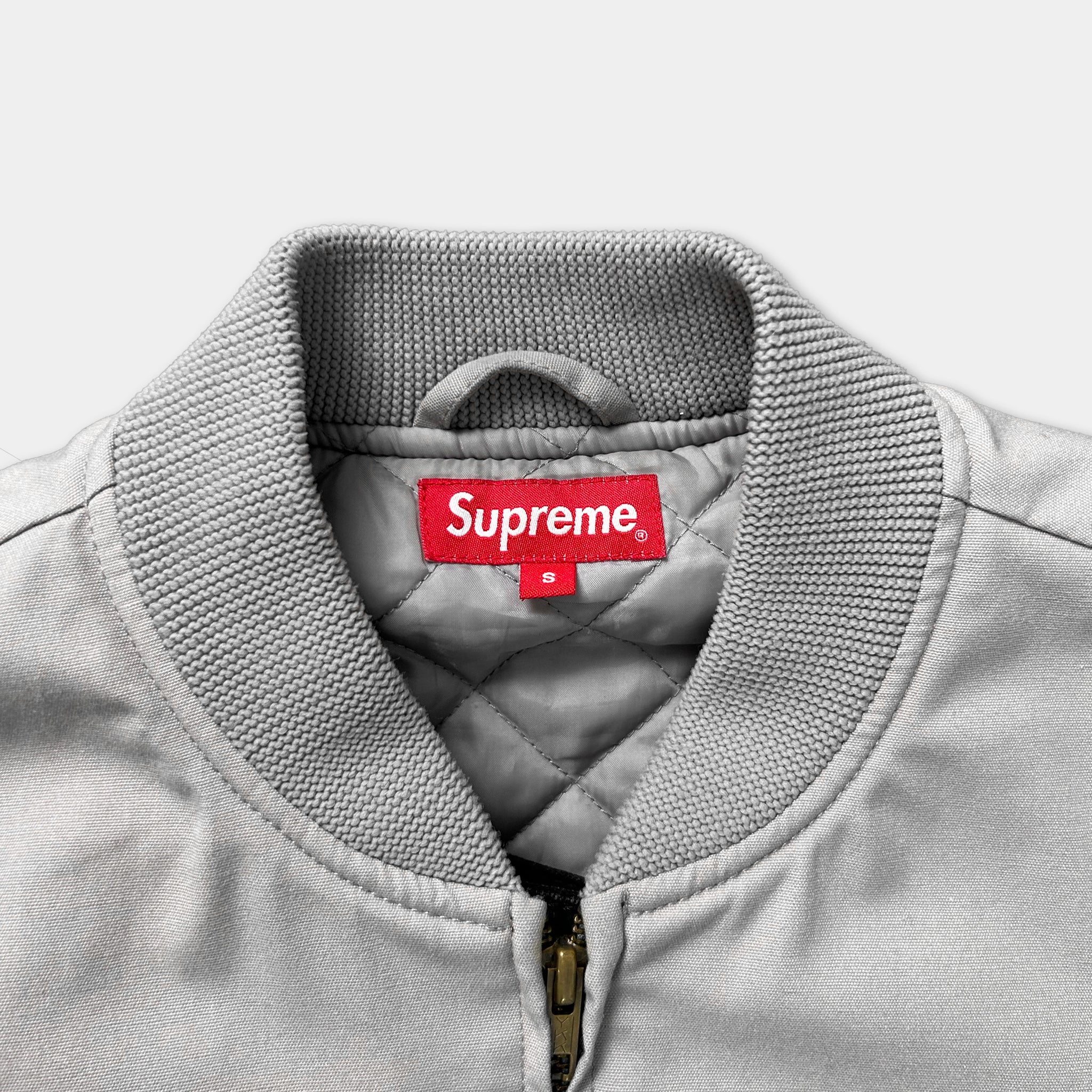 A gray Supreme jacket with a red label on the collar.