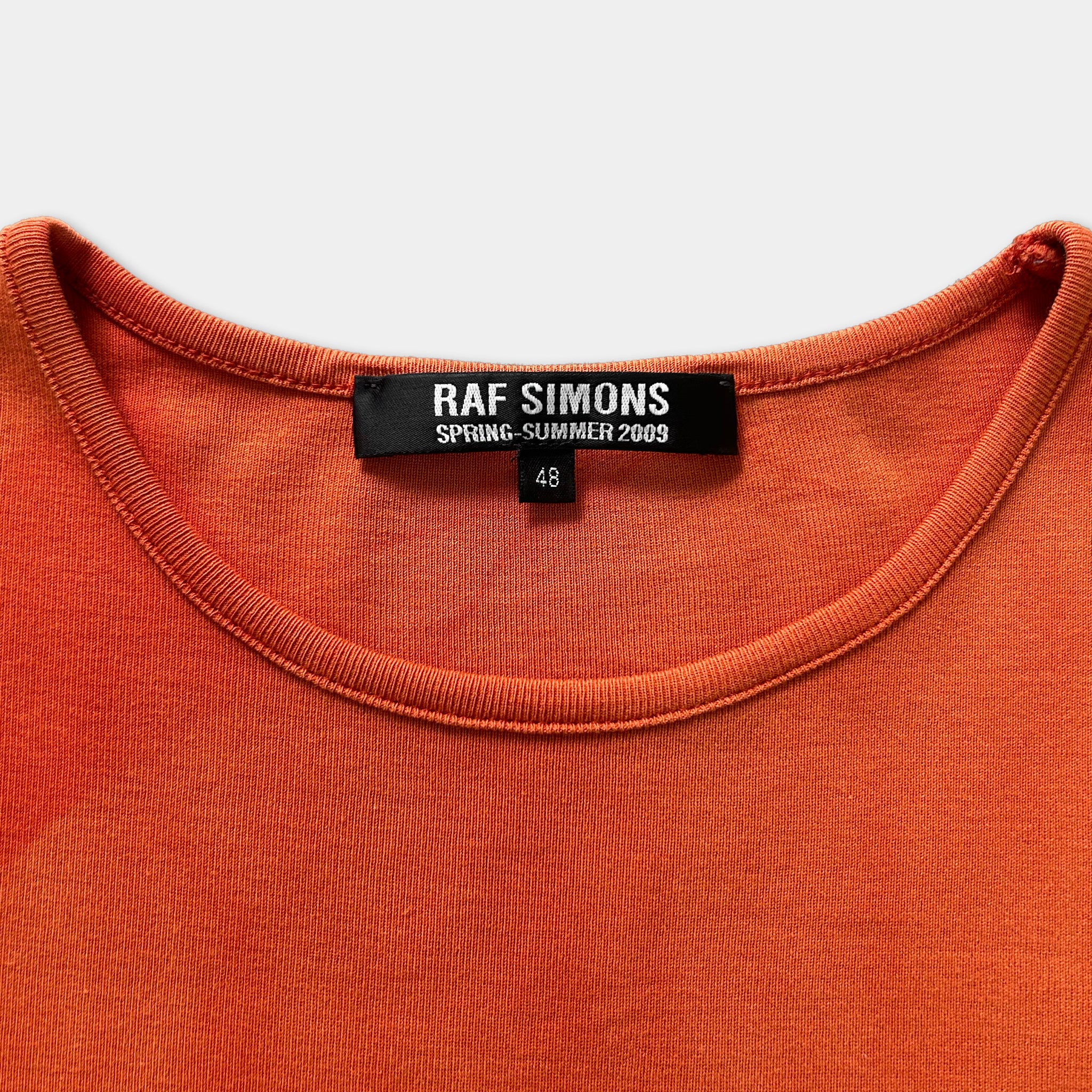 a close-up view of a bright orange t-shirt with a label that reads "RAF SIMONS SPRING- SUMMER 2009 48".