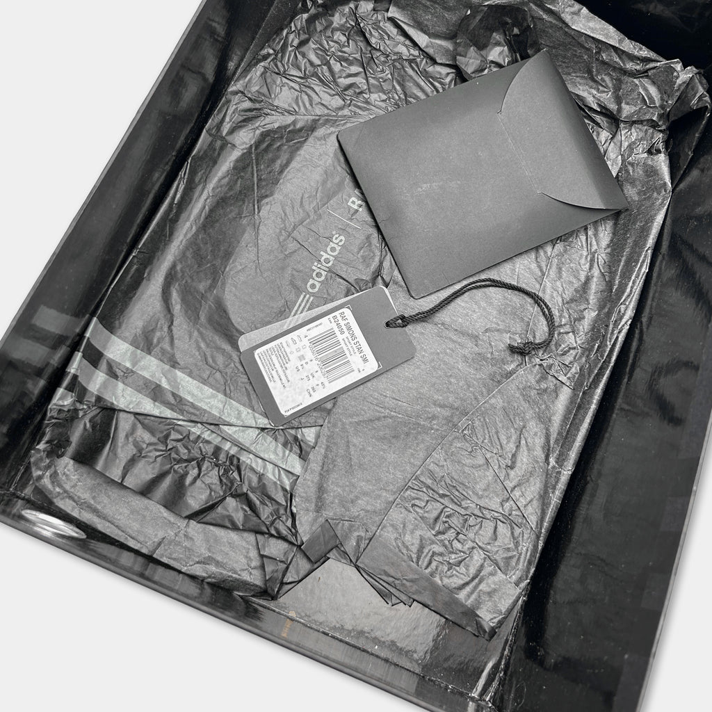 A black Adidas jacket is placed inside a black box, with a tag attached to it.