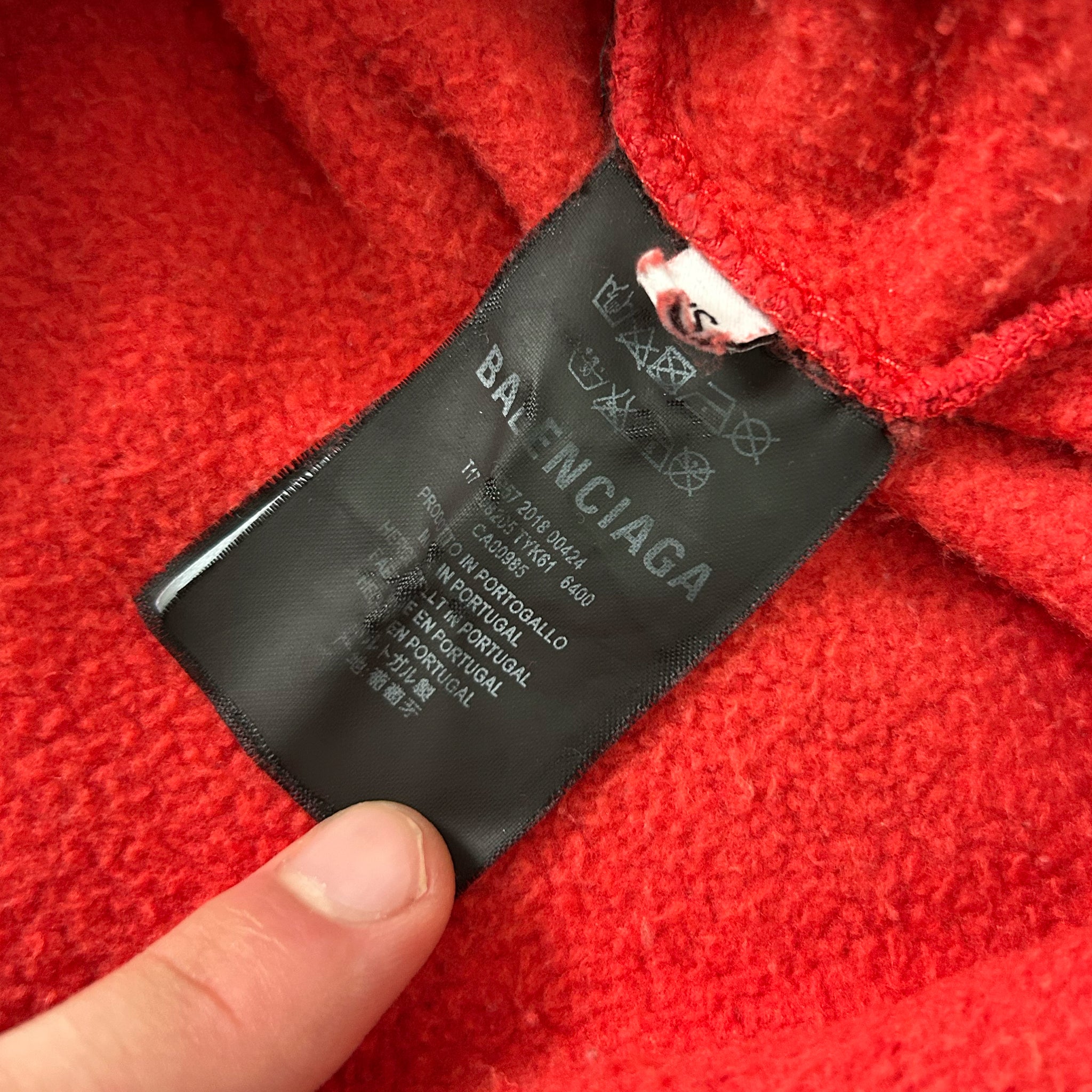 A close-up view of a red fabric, possibly a sweater or garment, with a black label attached to it. The label appears to be a garment label from the Balenciaga brand, indicating the brand name and possibly other details about the product.