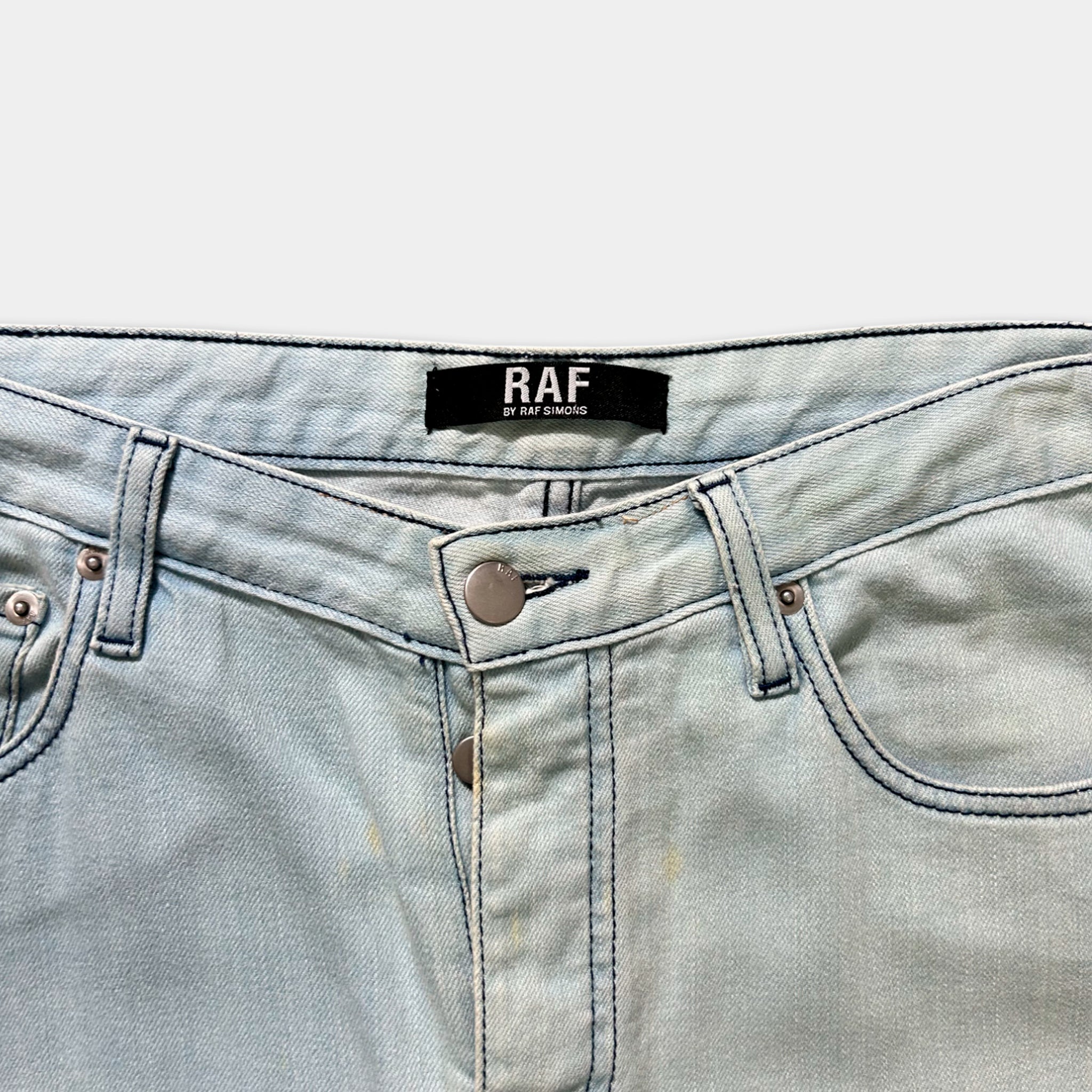 A pair of light blue denim jeans with a label that reads "RAF" and "BY RAFAEL" printed on the waistband.