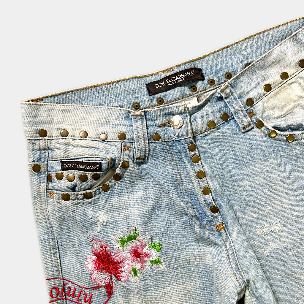A pair of blue denim jeans with floral embroidery and metal studs on the pockets.