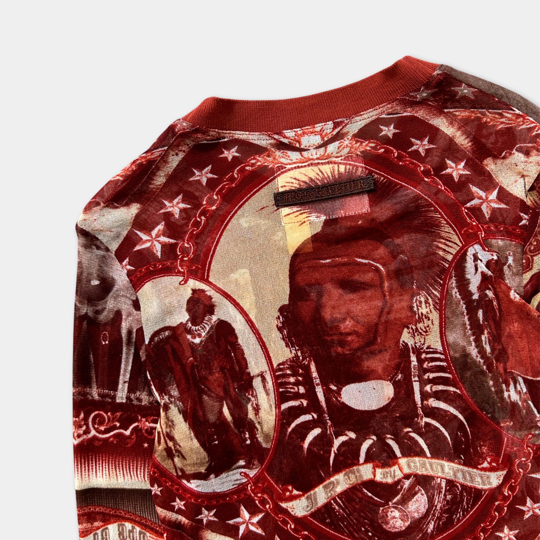 The image is a close-up of a red and gold patterned fabric, featuring a central image of a person with a large headdress and a necklace.