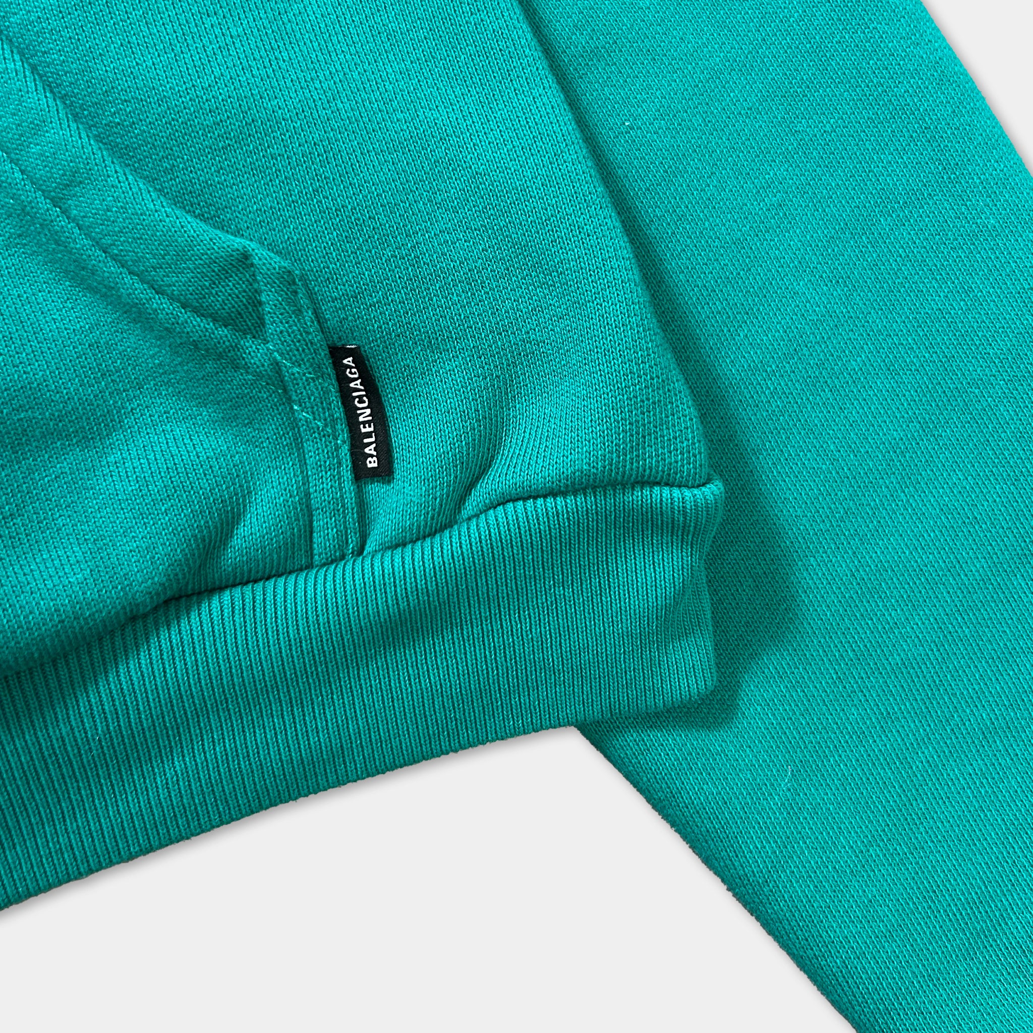 A close-up view of a turquoise-colored hoodie or sweatshirt, with a small black label or tag attached to the fabric.
